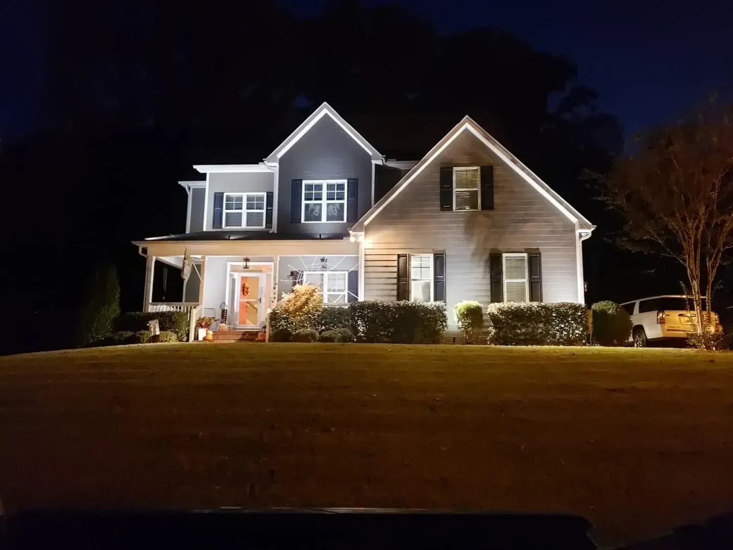 Two-story house illuminated at night. Gray siding with white trim. Lights shine on the porch and bushes. A vehicle is parked in the driveway.