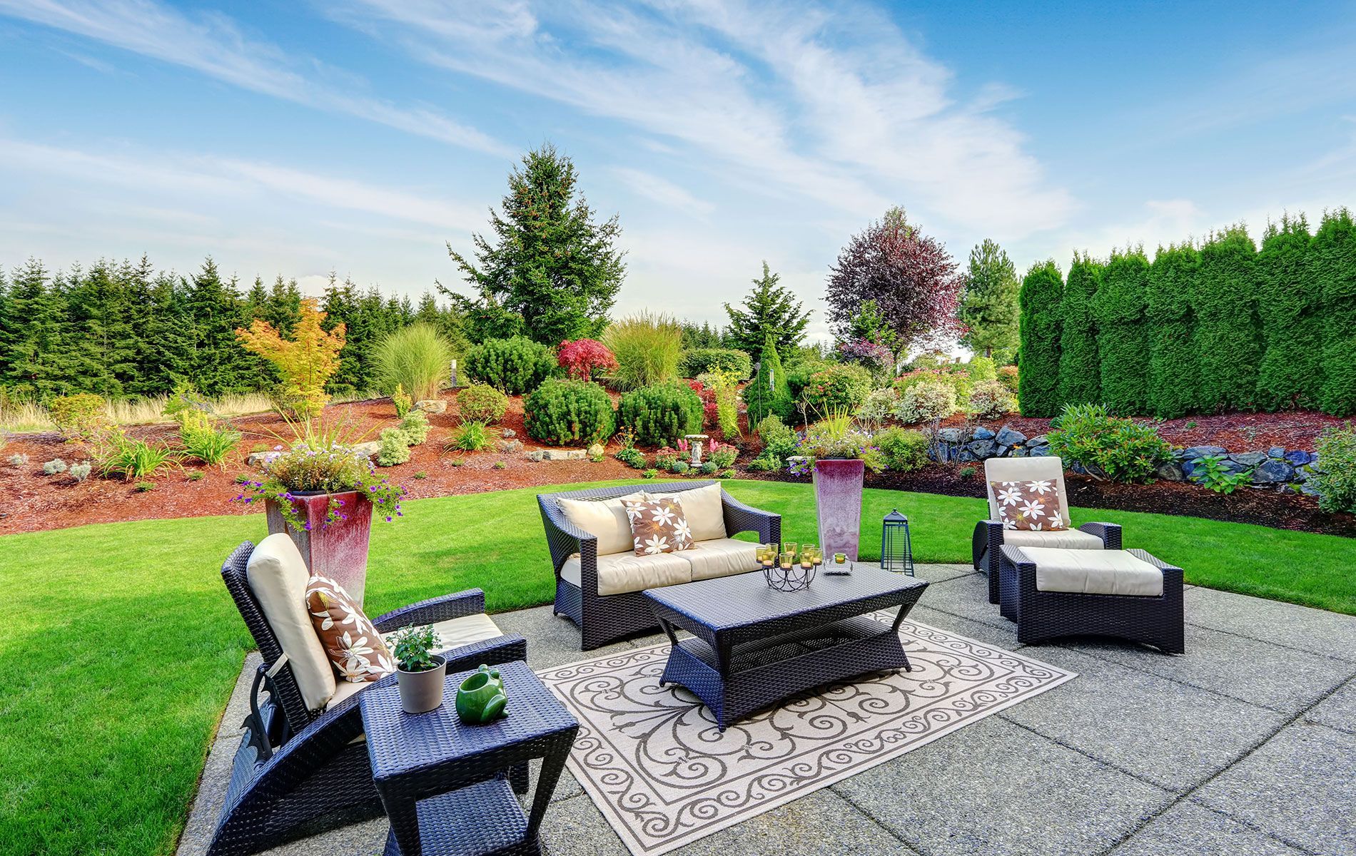 Patio with wicker furniture on a patterned rug, surrounded by green lawn and garden under a blue sky.