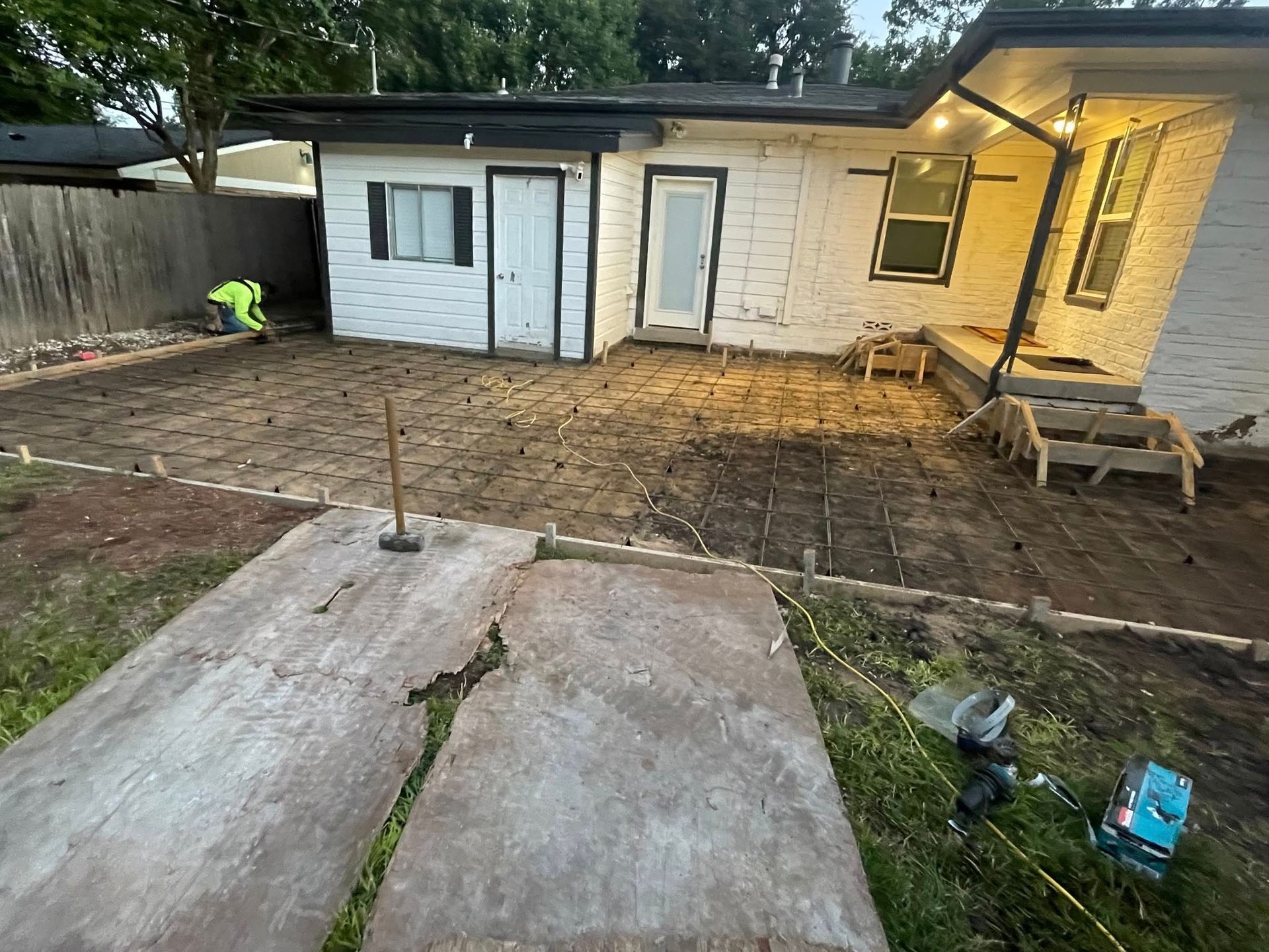 Backyard concrete work in progress; white house, dirt, and a worker in safety vest.