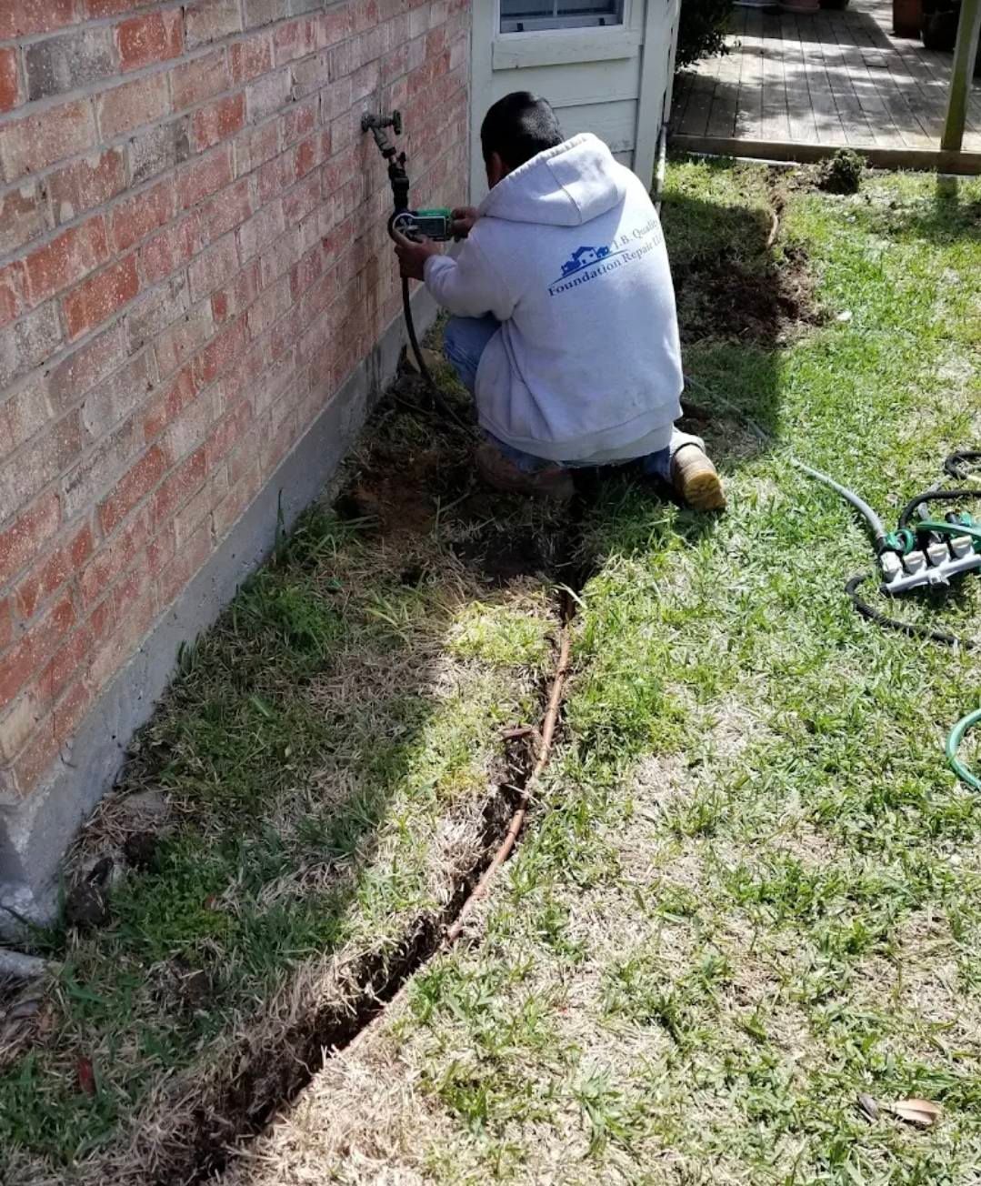 Person kneeling by brick wall, working on a pipe in a shallow trench in the grass.