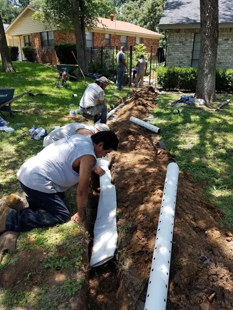 Workers install drainage pipes in a trench in a grassy yard, near houses.