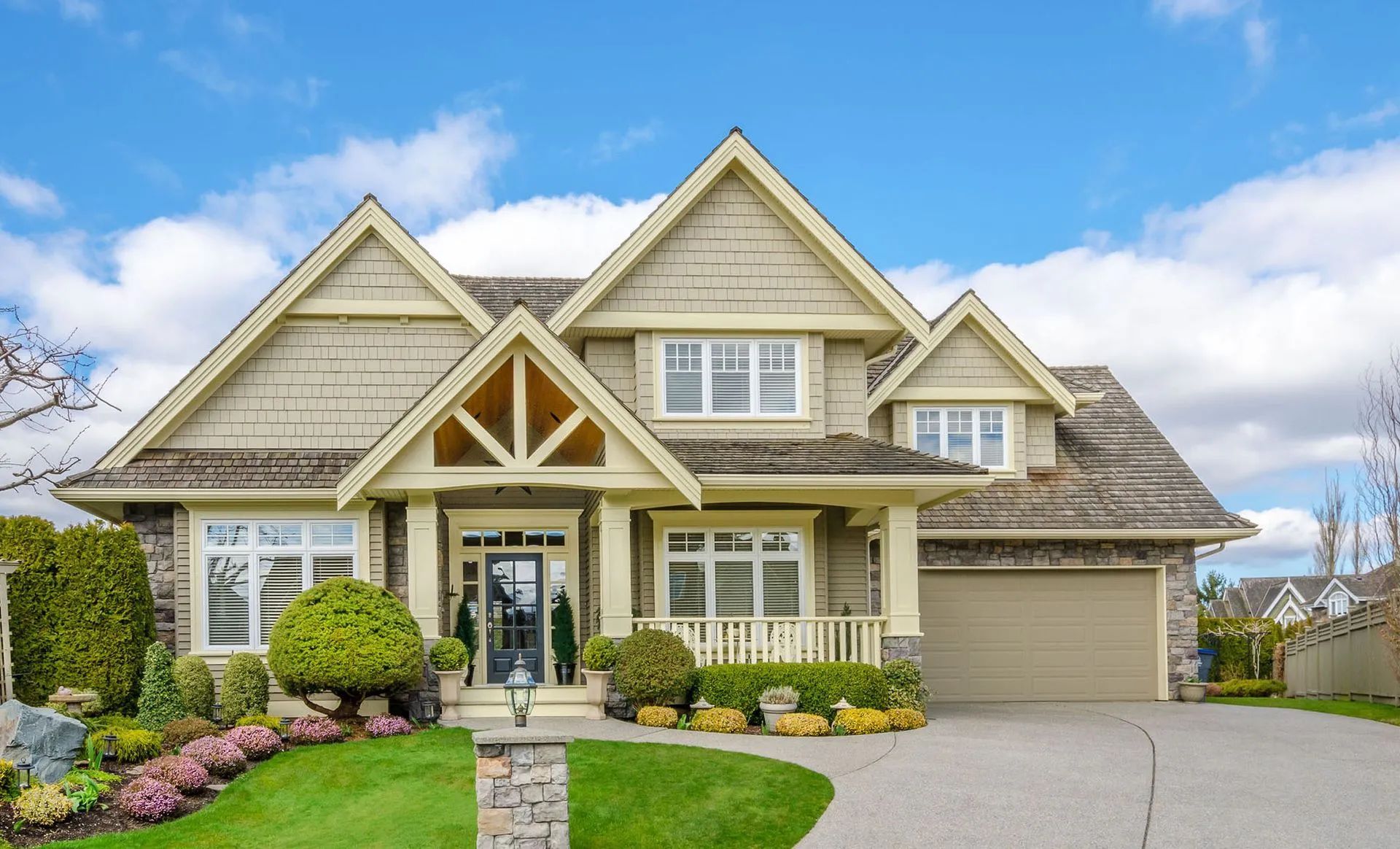 Two-story beige house with a stone porch, garage, and blue sky. Green lawn and shrubs in front.