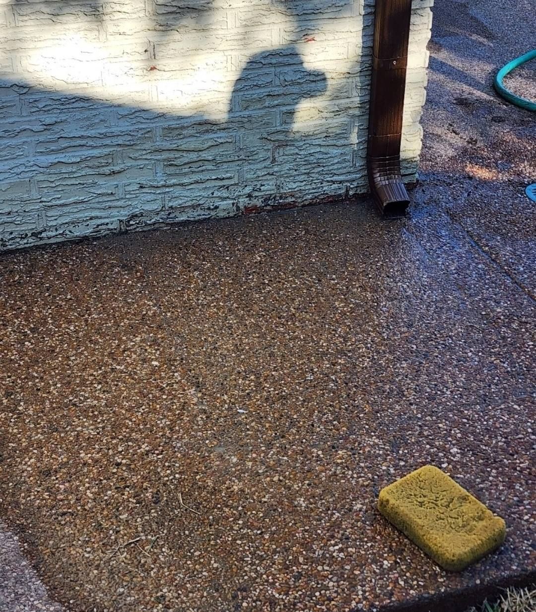 Wet, speckled concrete patio with a yellow sponge. A shadow is cast on a textured wall.
