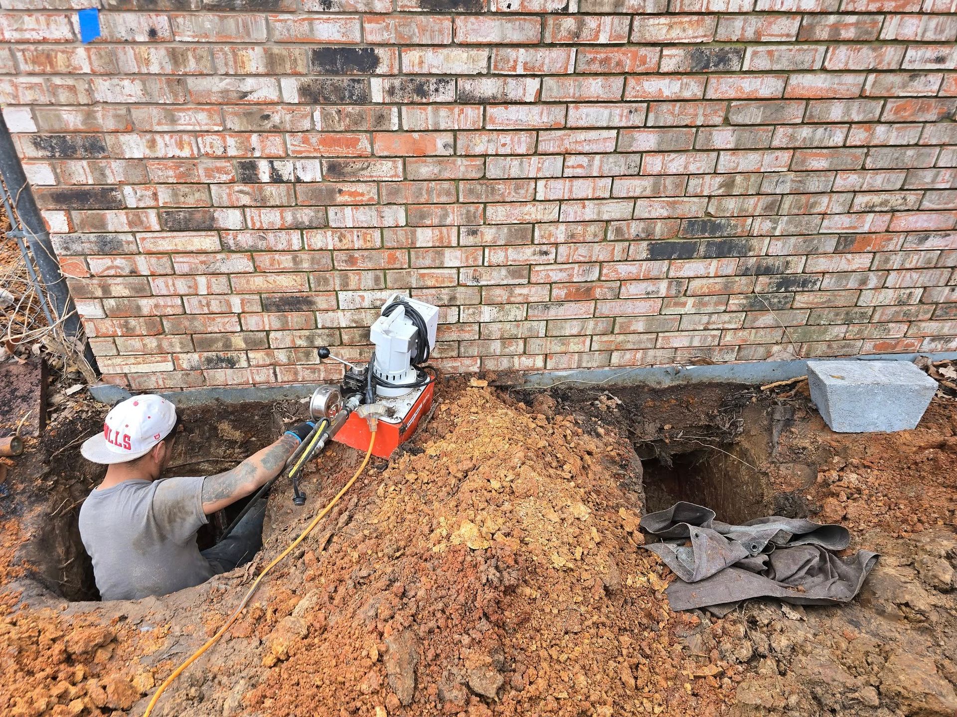 Man operating a jackhammer next to a brick foundation, soil excavated.