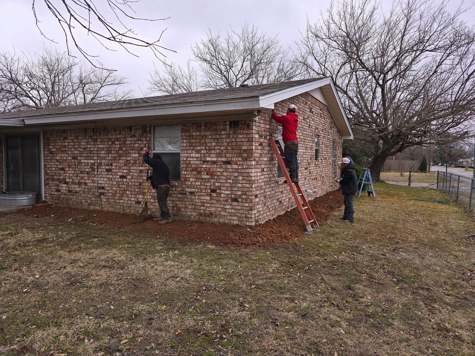 People working on the brick exterior of a house. One person on a ladder, two others on the ground. Overcast day.