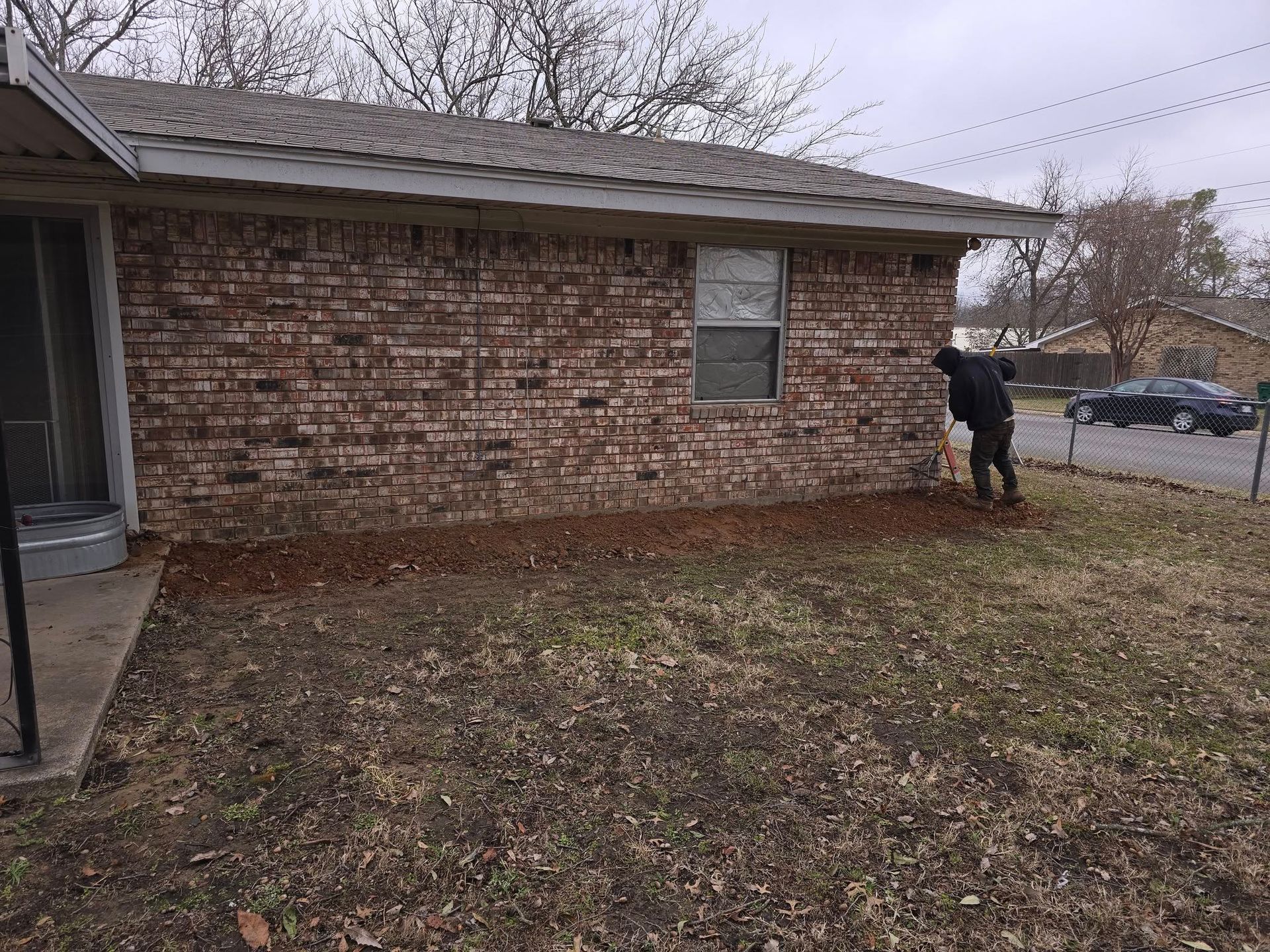 Person raking fallen leaves in front of a brick house with a brown roof and window on a cloudy day.