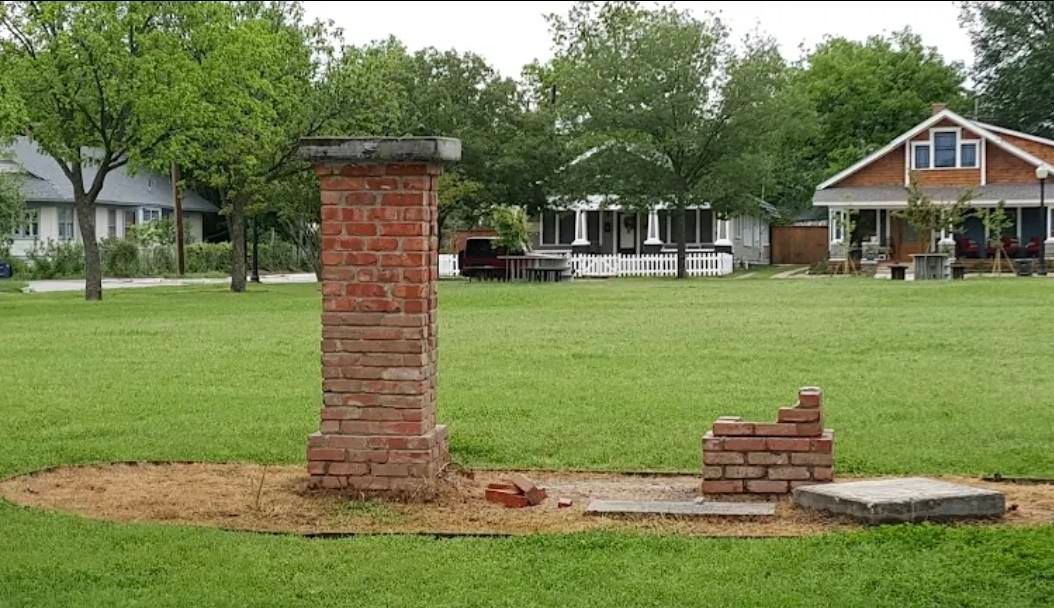 Brick chimney remnant and small brick structure on grassy lawn; houses and trees in background.