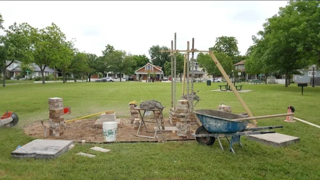 Construction site in a grassy park; wheelbarrow, tools, and partial structure in progress, residential houses in the background.
