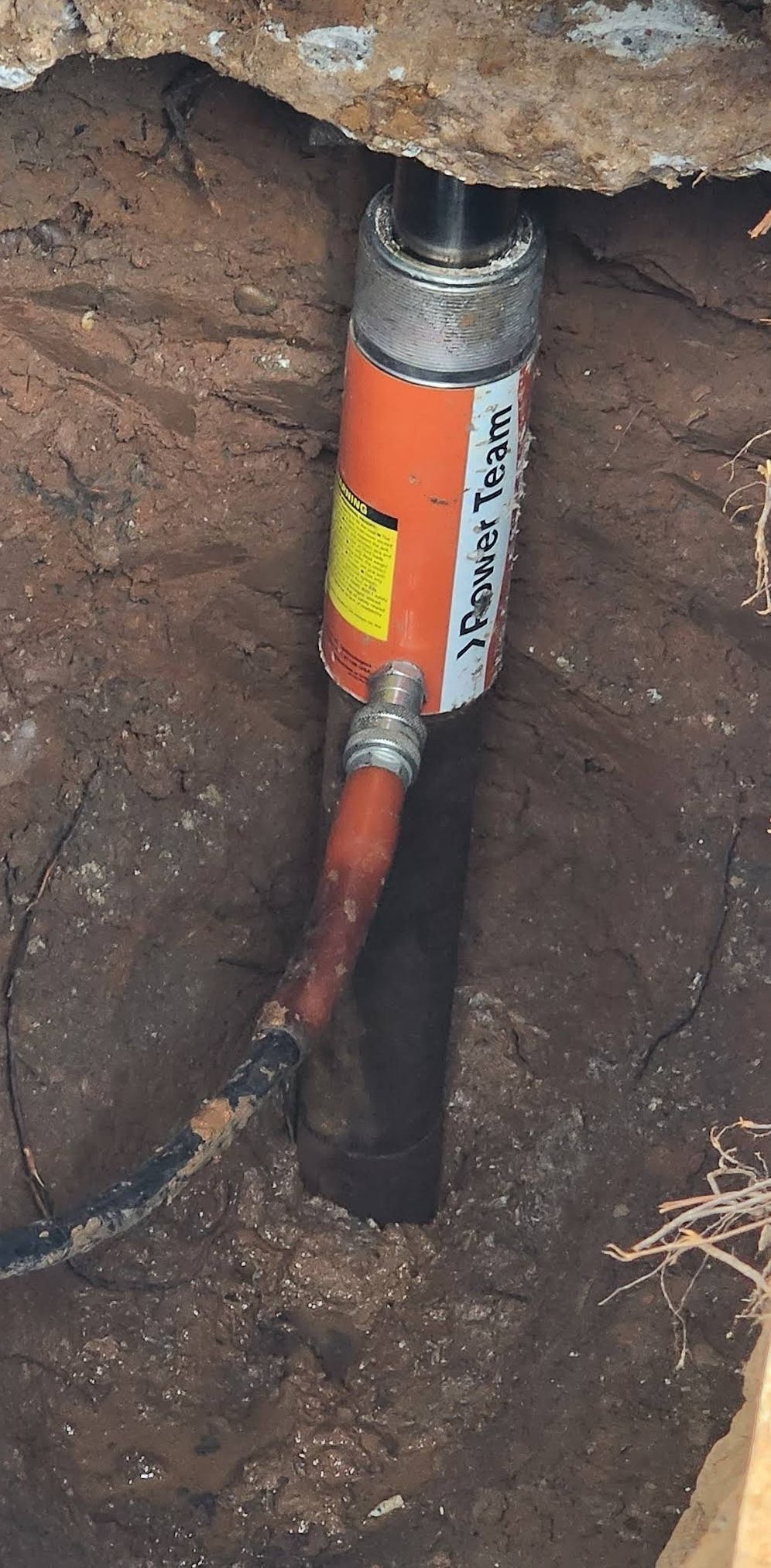 An orange hydraulic jack is set in a dirt trench, supporting something above.