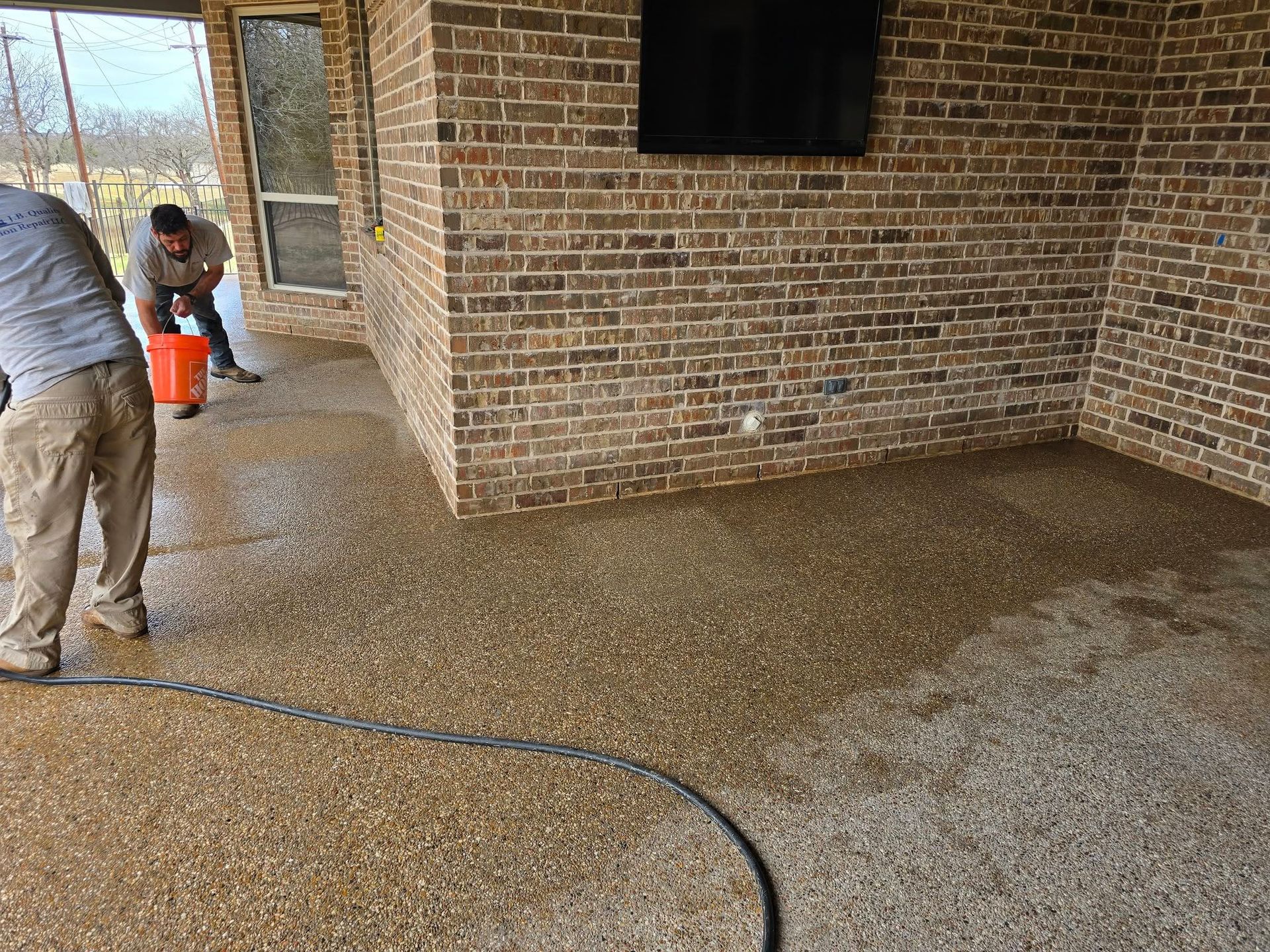 Two people cleaning a textured concrete patio under a brick-walled covered porch.