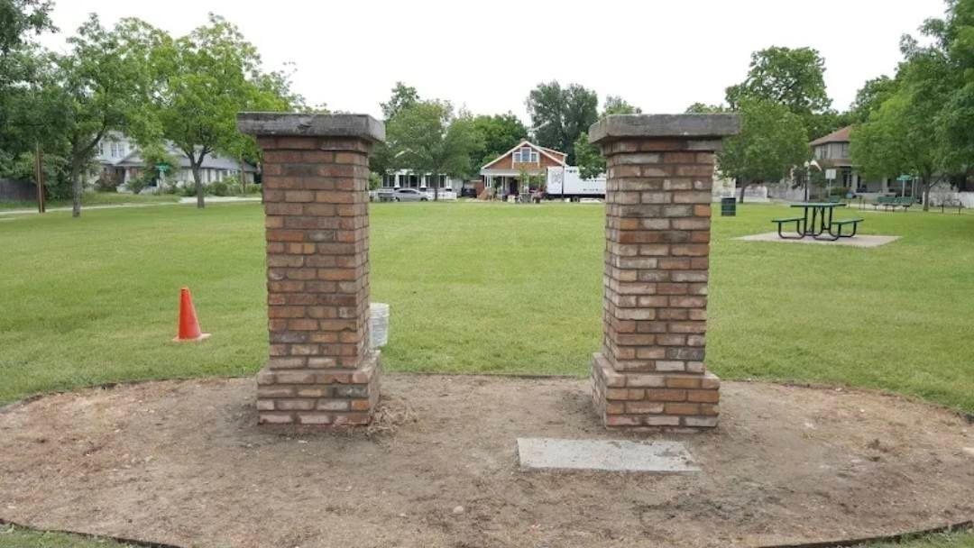 Two brick columns with concrete tops in a grassy park setting; picnic table and trees in the background.