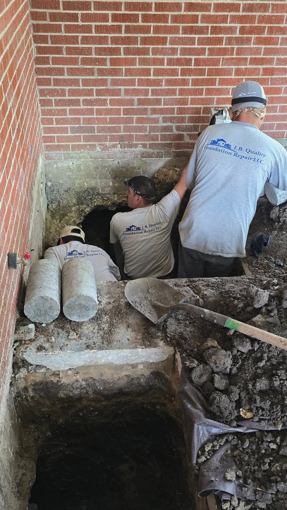 Three construction workers excavating near a brick wall, two are in the hole.
