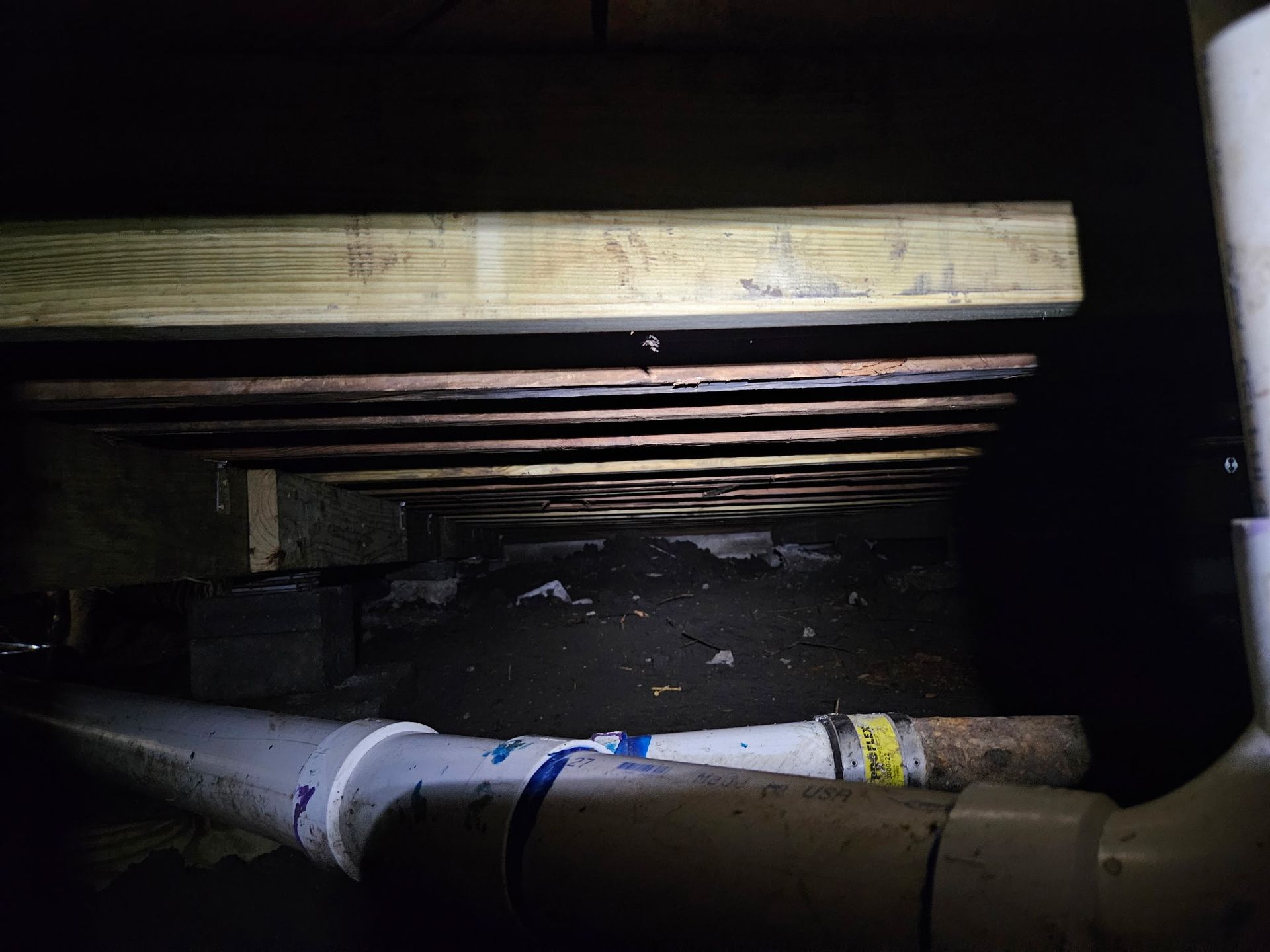 View from below of a crawl space with joists, wooden beams, plumbing, and dark soil.