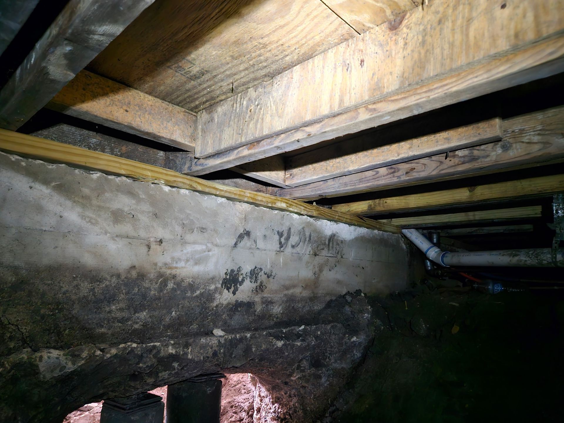 View of a crawl space under a house, showing concrete foundation wall, wooden beams and framing.