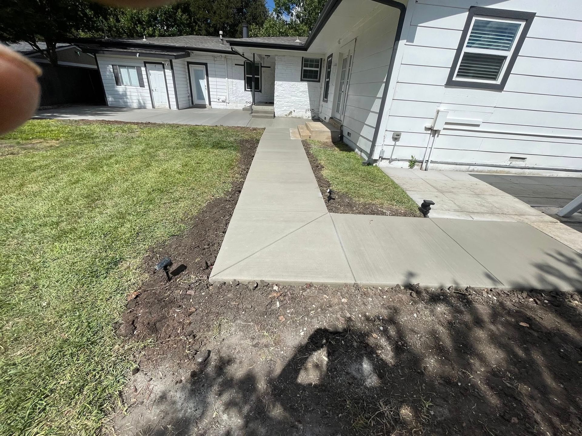 A newly poured concrete walkway leads to a white building with black doors. Green grass and landscaping surround.