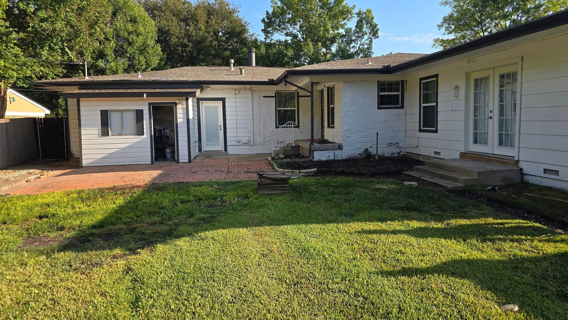 White house exterior with overgrown grass, red gravel, and trees in the background.