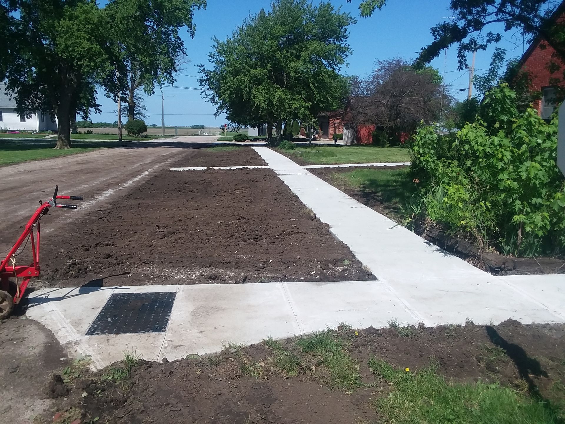 A red plow sits in the dirt next to a concrete walkway