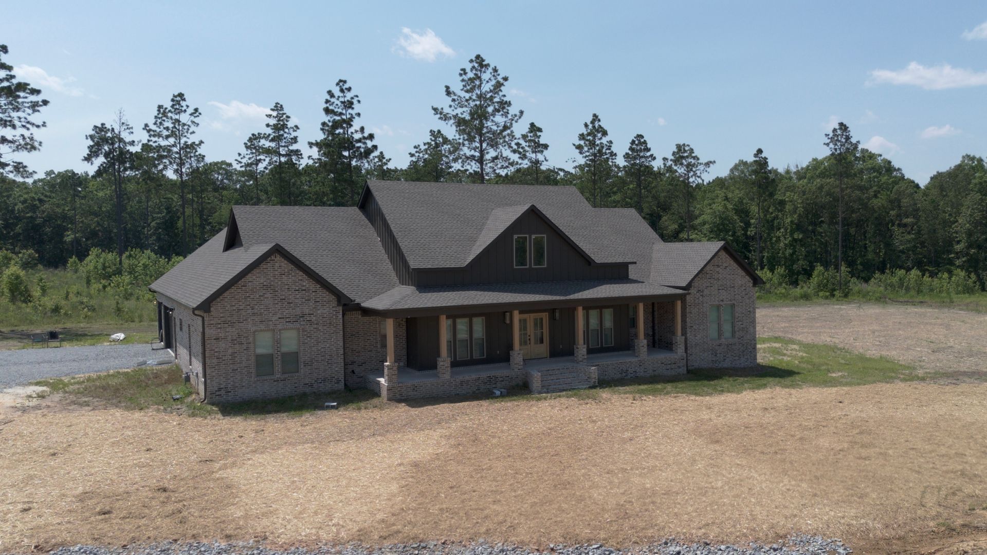 Brick house with dark roof and porch, set in a clearing near a forest under a blue sky.