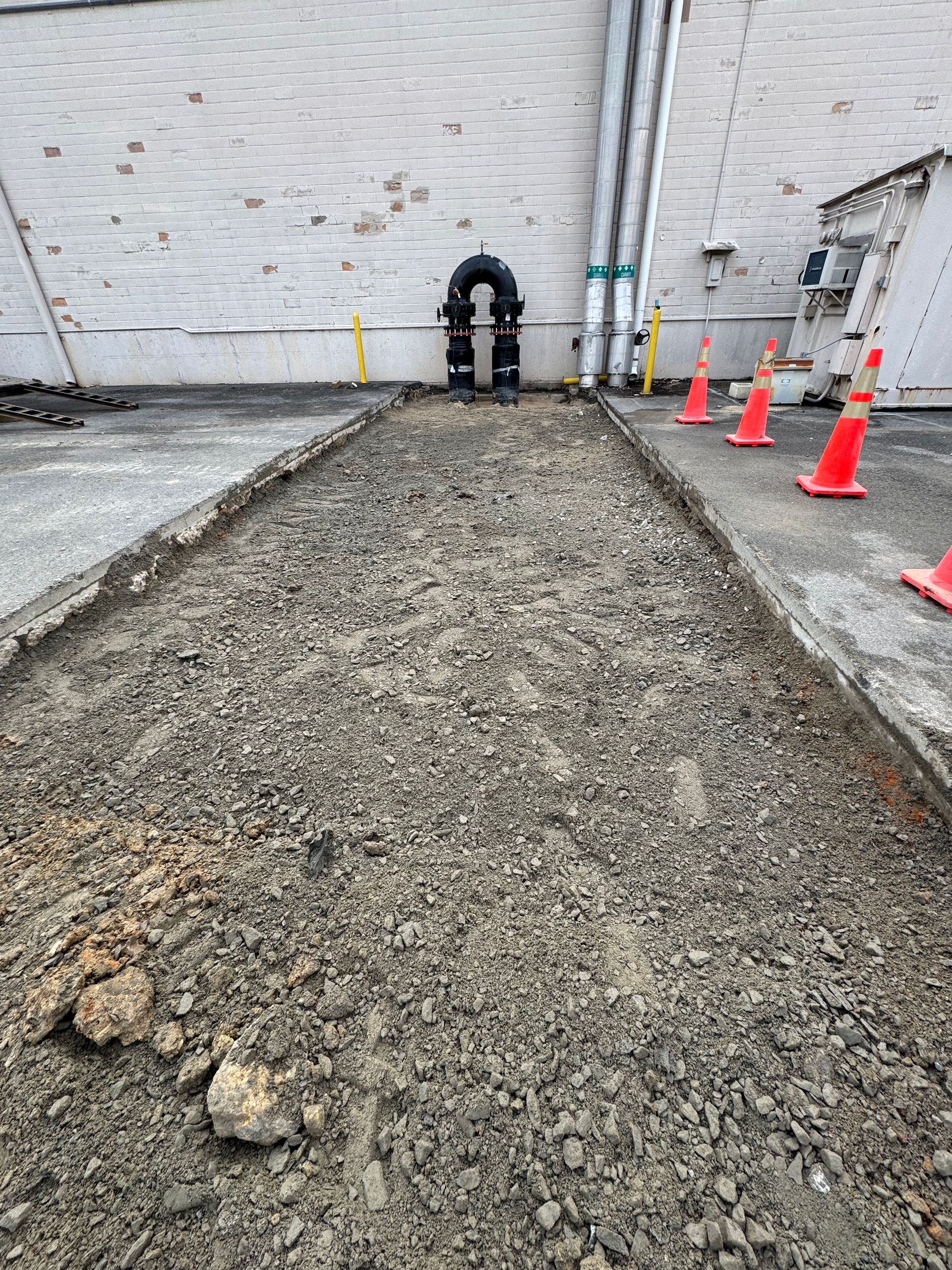 Outdoor area with excavated strip; gray gravel, orange cones, and pipes against white brick building.