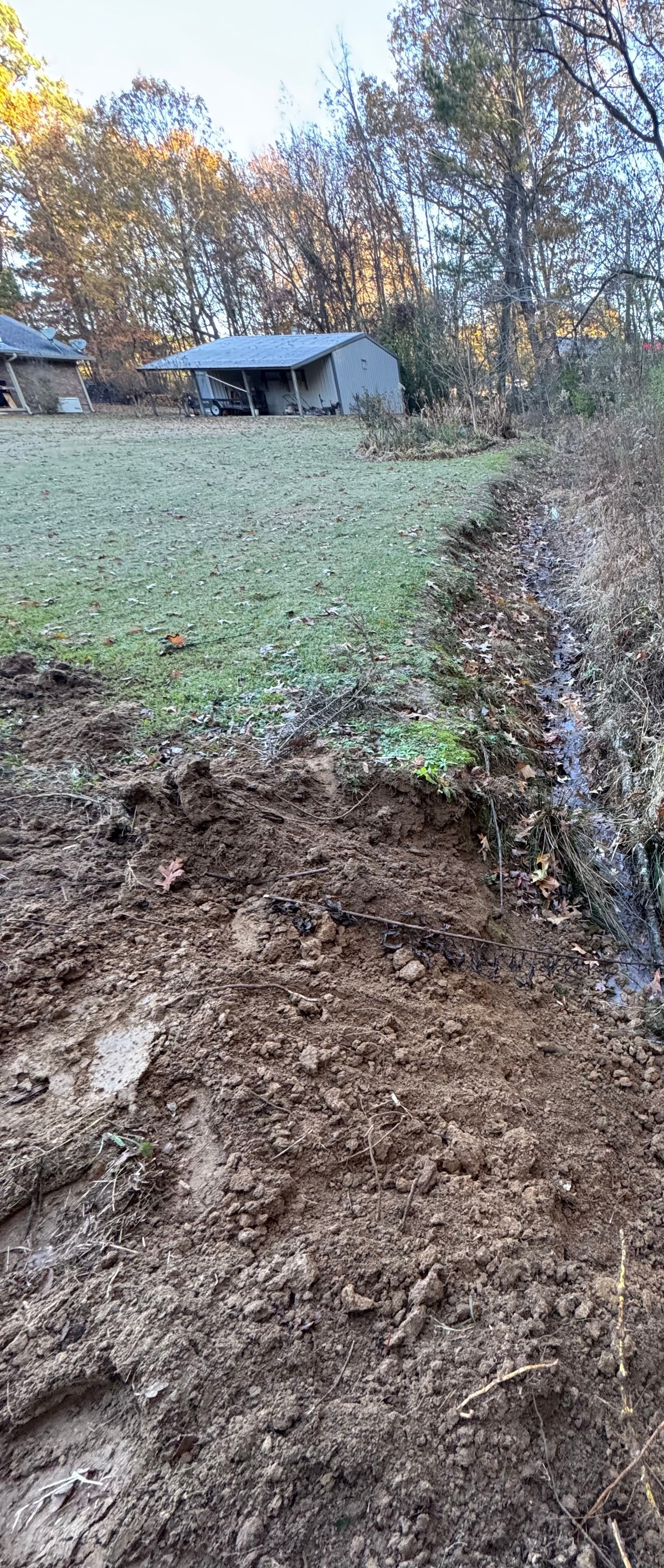Dirt mound and muddy trench next to a grassy field with a shed and trees in the background.