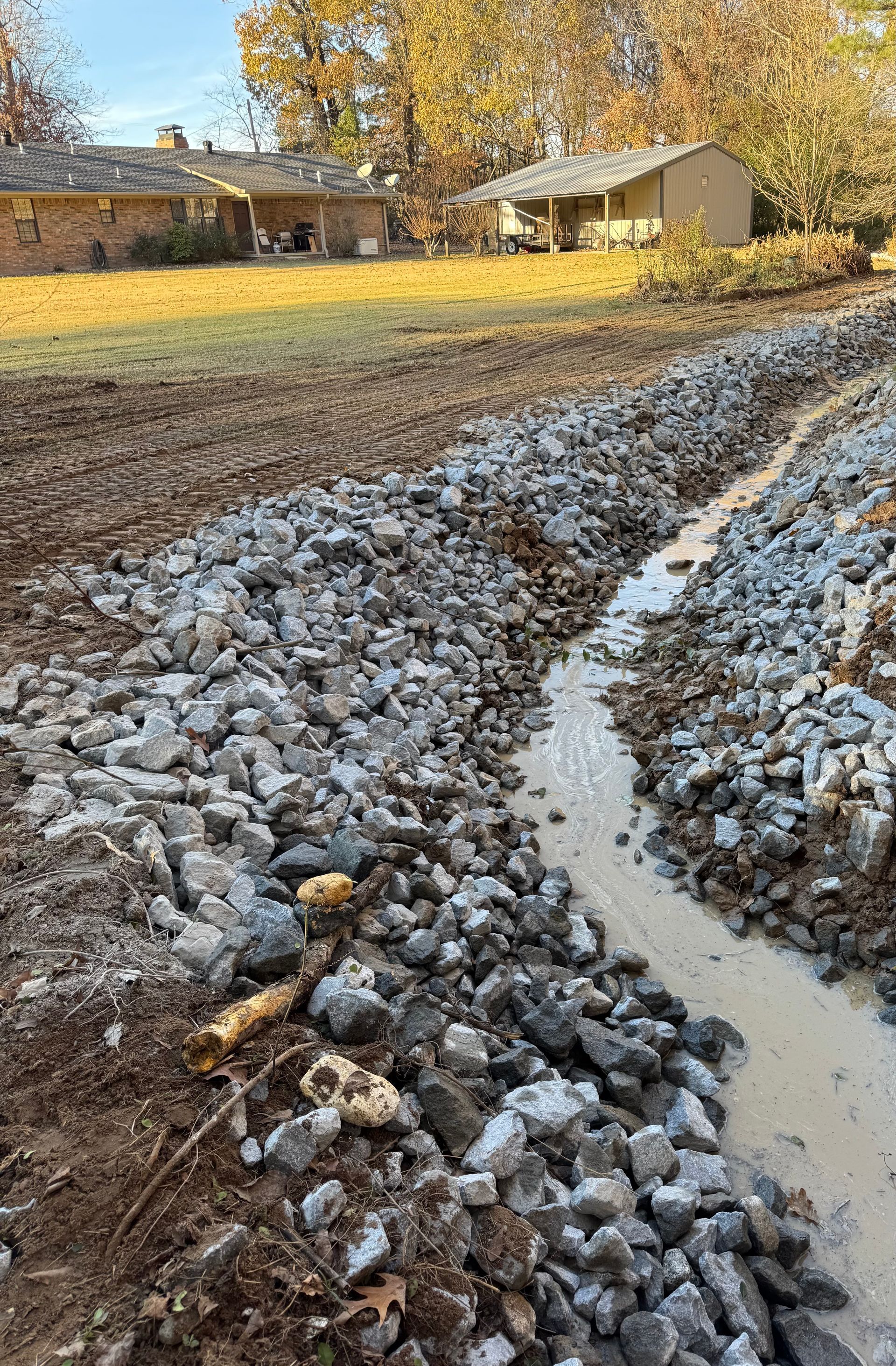 A rocky drainage ditch with muddy water, next to a field and houses.