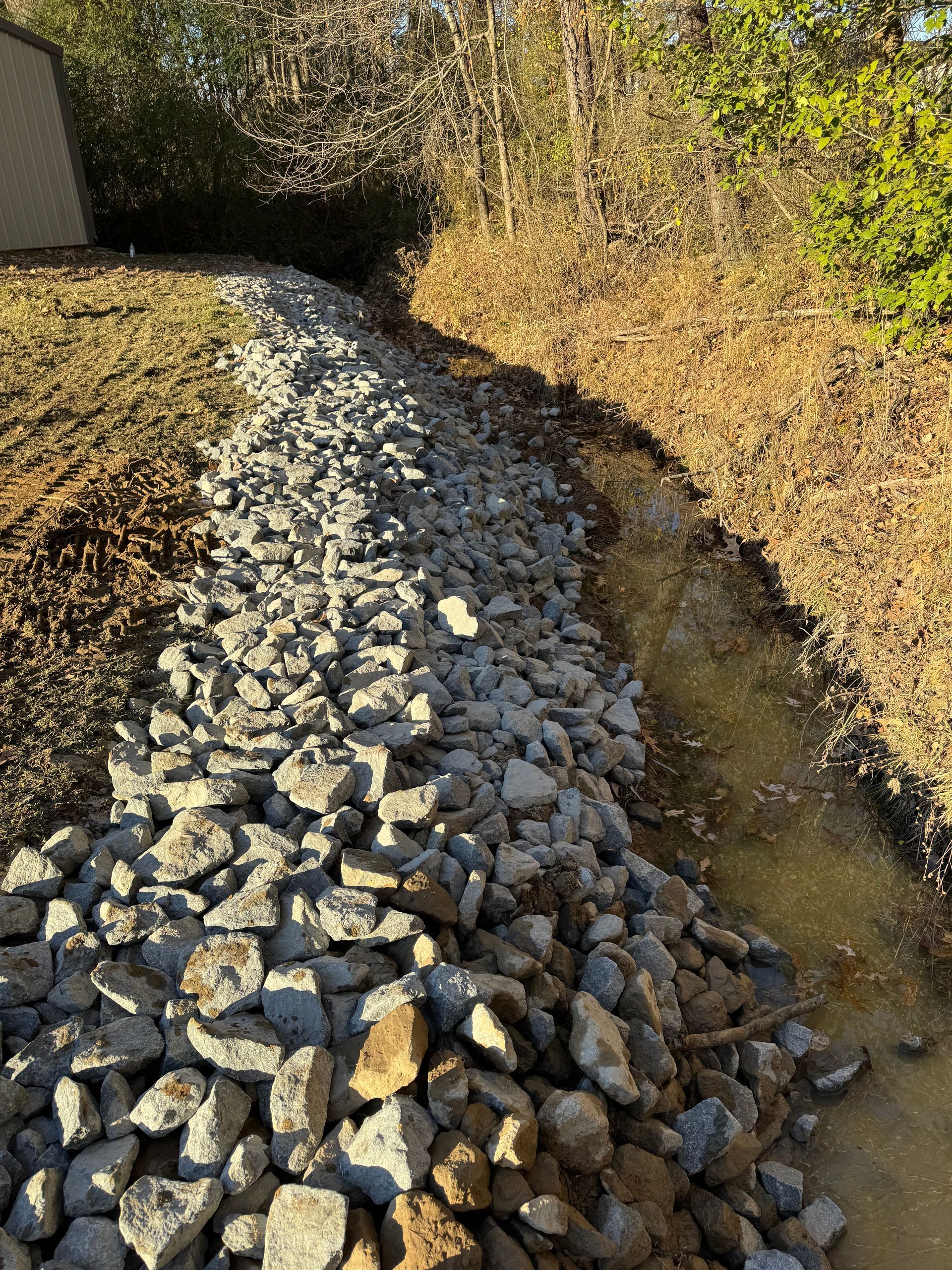 Gravel-lined ditch runs alongside a grassy embankment and a wooded area. Sunny daytime.