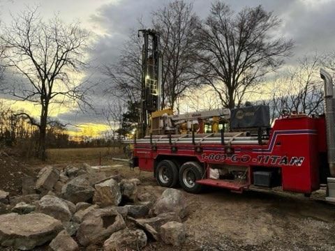 Red tow truck on a dirt path beside a rock pile at sunset, with bare trees and a cloudy sky.