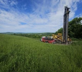 Red drilling rig beside a truck in a green field under a cloudy blue sky