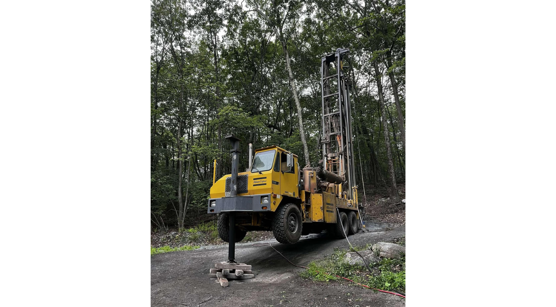 Yellow drilling rig in a wooded area, with a person working beside it on rough ground.