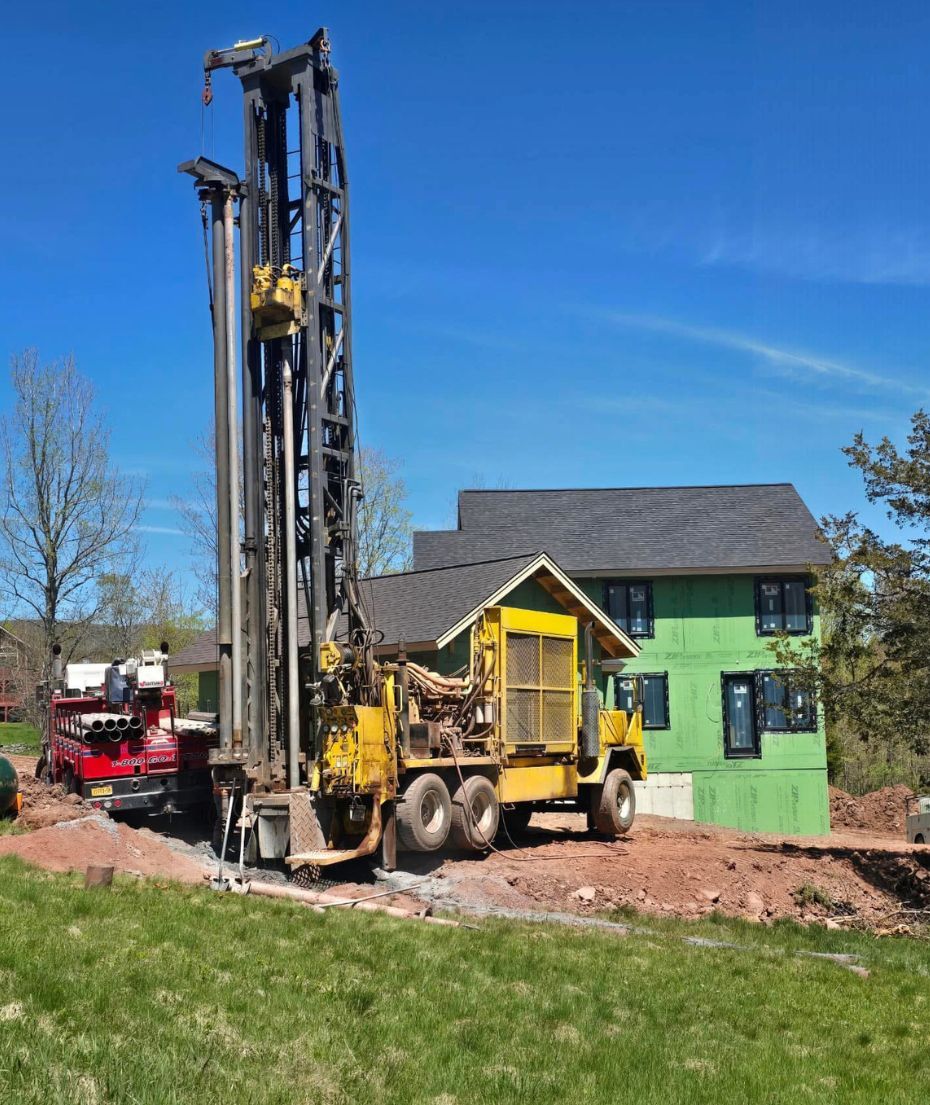 Yellow drilling rig, red support truck, near a house with green siding, on a sunny day.