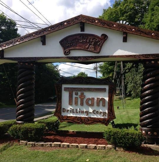 Sign for Titan Drilling Corp, brown and white, with ornamental columns and scrollwork; green bushes below.