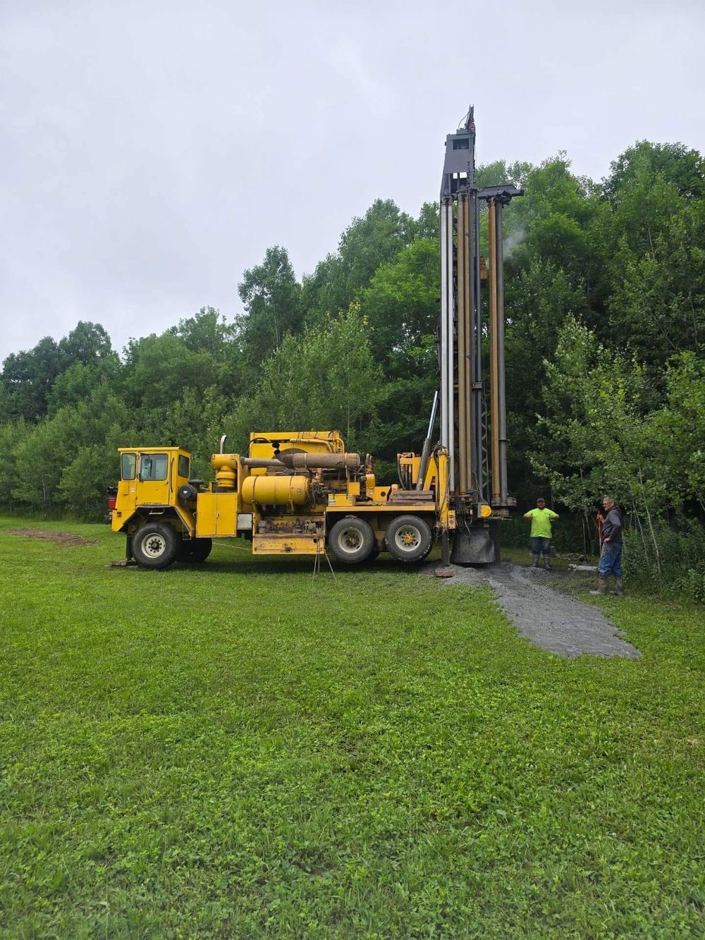 Yellow drilling rig in a grassy field, near trees. Two people are working near the rig under overcast skies.