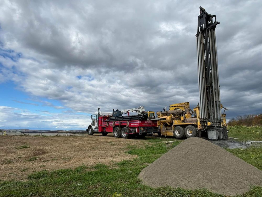 A red and yellow drilling rig on a field under a cloudy sky. A pile of sand sits near the rig.