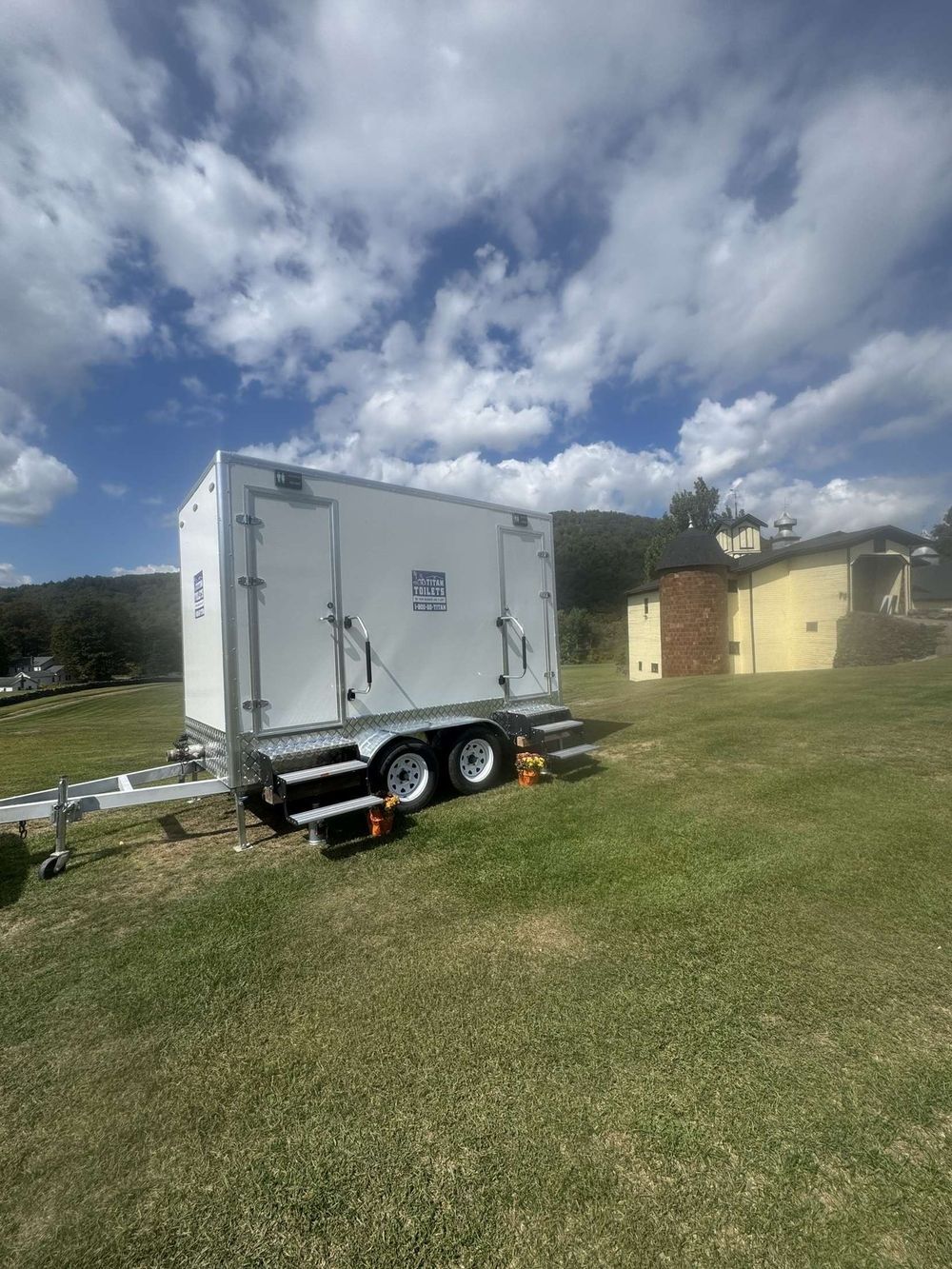 White portable restroom trailer on a grassy field; cloudy sky, small yellow building in the background.