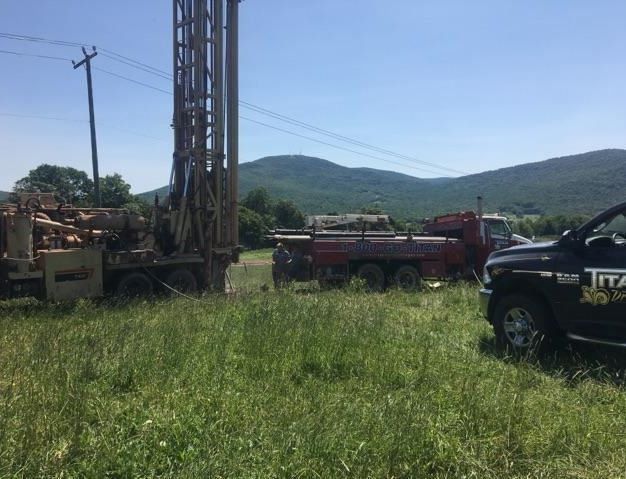 Drilling rig and water truck on a grassy field with a mountain backdrop under a bright sky.