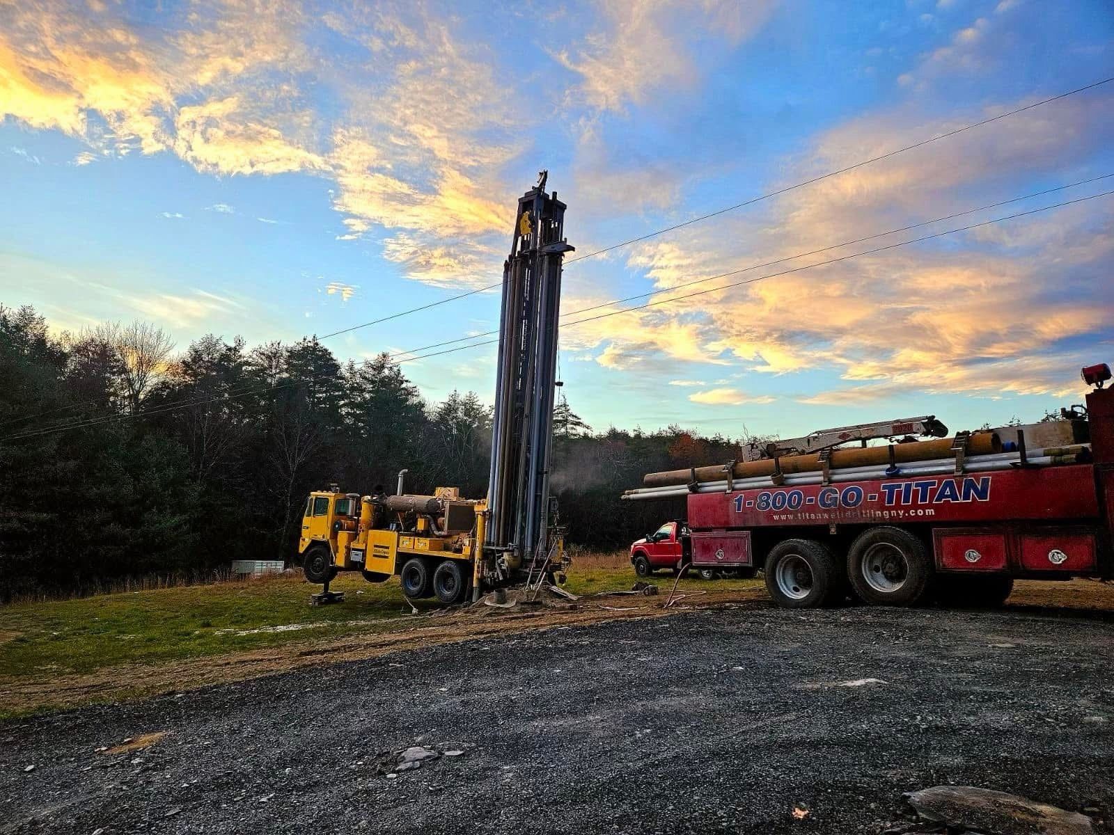 Yellow drilling rig and red truck at a construction site under a cloudy sunset sky