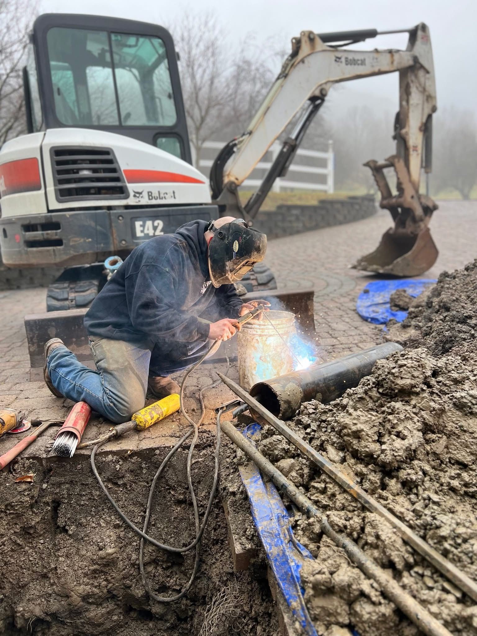 Construction worker welding beside excavator at a muddy worksite with exposed pipes and blue tarp