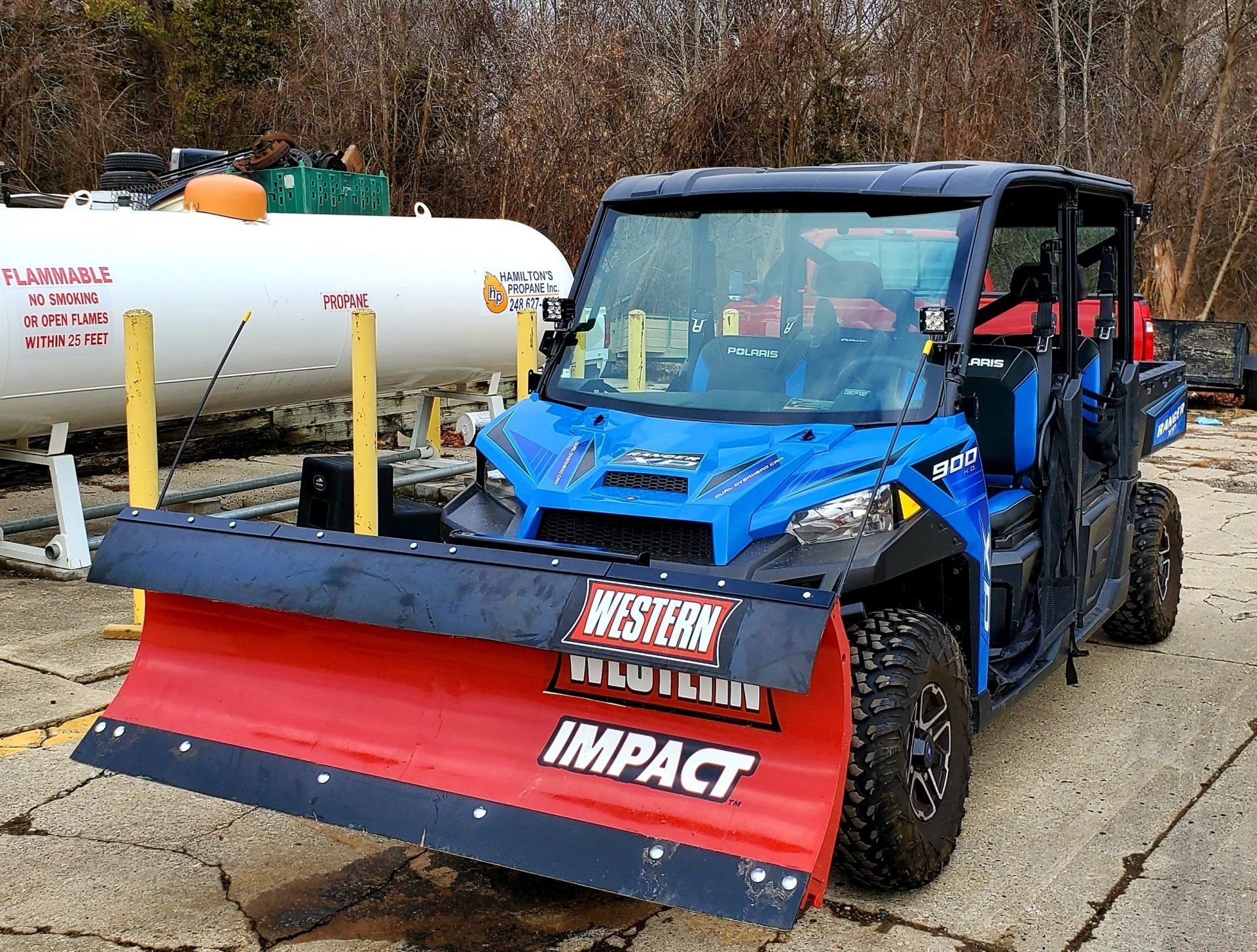 Blue utility vehicle with a red snowplow blade parked outdoors beside a white tanker