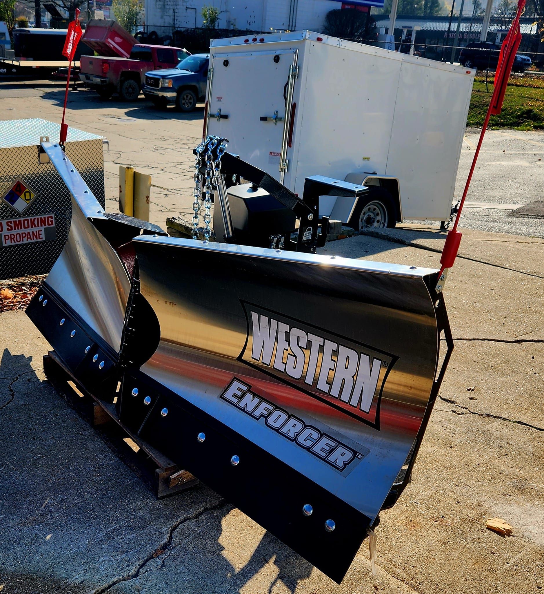 Black snowplow blade with white lettering displayed on a lot, with a truck and equipment in the background