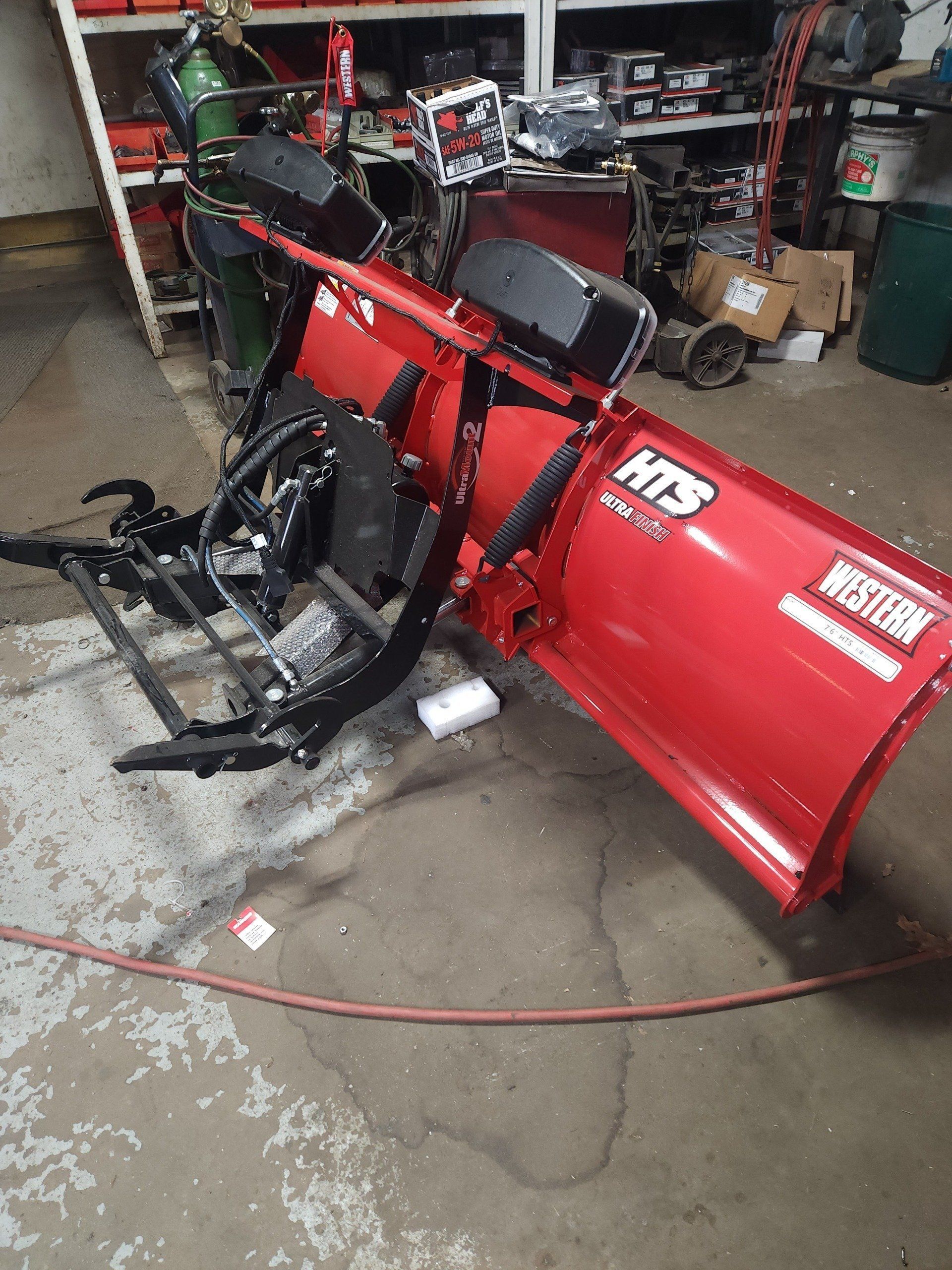 Red snowplow blade attached to a black mounting frame in a garage with tools and equipment.