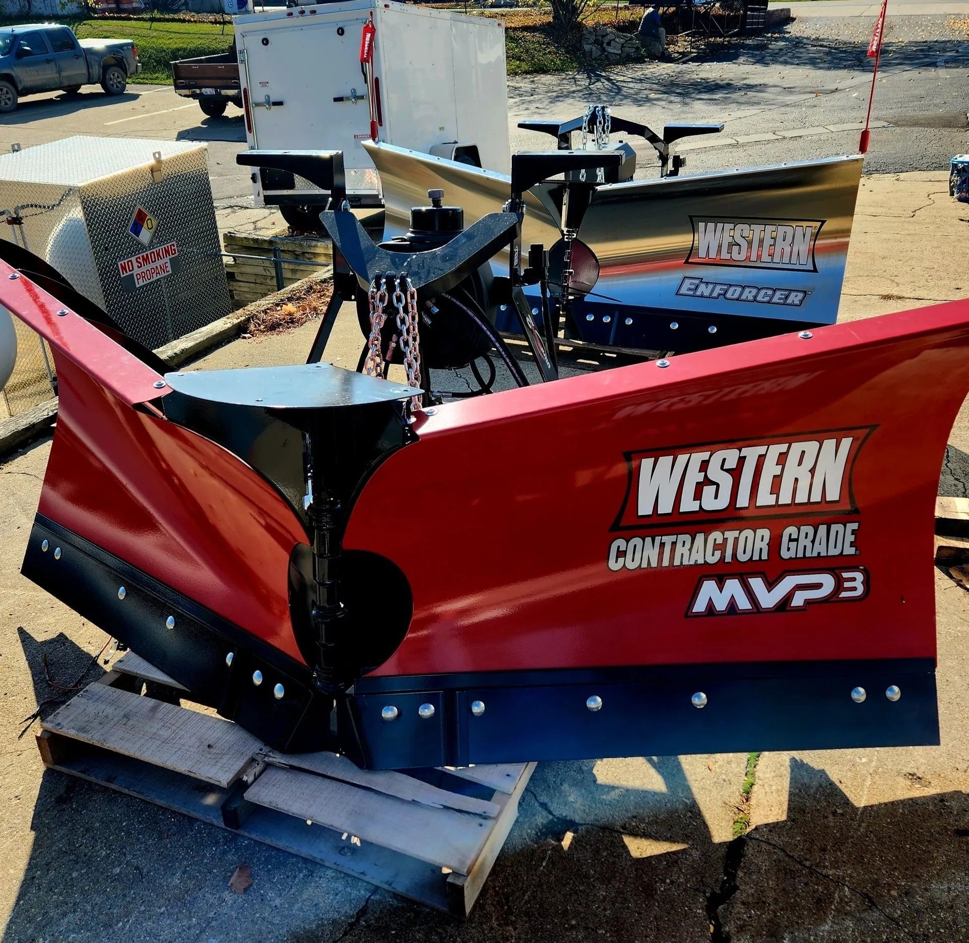 Red Western Contracting Grade snow plow attachment on pavement, with blue truck and construction equipment in background