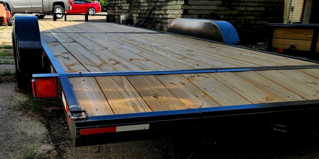 Black flatbed trailer parked outdoors on gravel, with wood deck and blue fenders