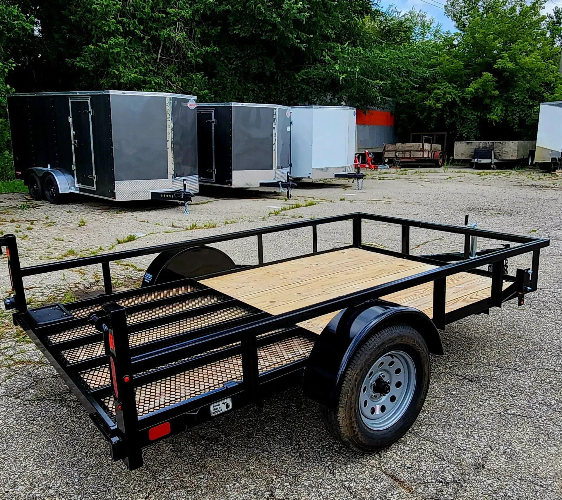 Black utility trailer with mesh sides and wooden floor parked on gravel outdoors.