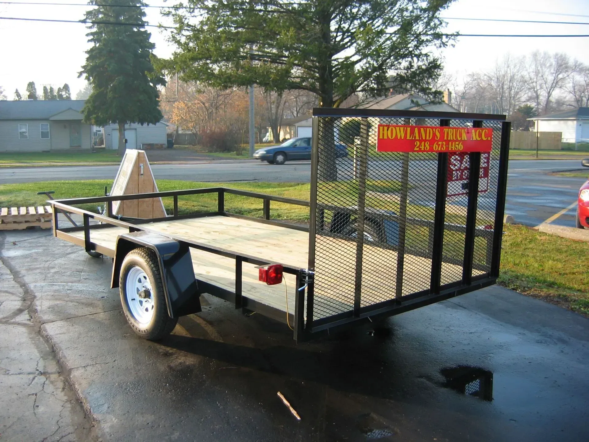Utility trailer with mesh ramp and yellow sale sign parked on wet pavement outdoors