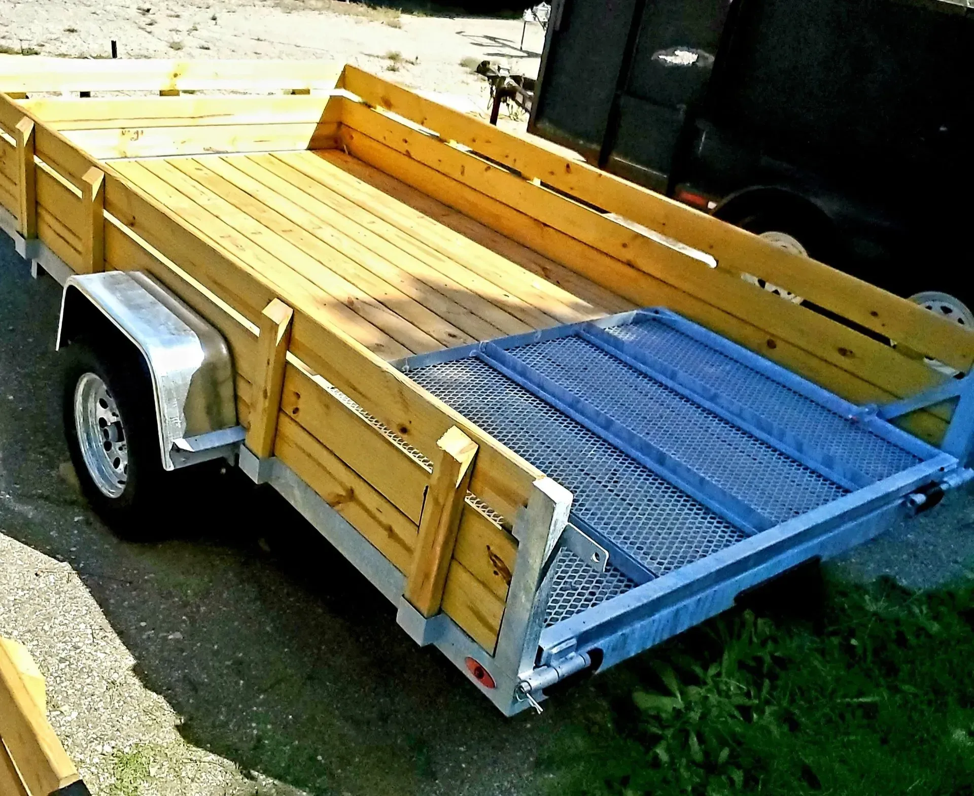 Small yellow utility trailer with wooden sides and a blue mesh ramp parked outdoors.