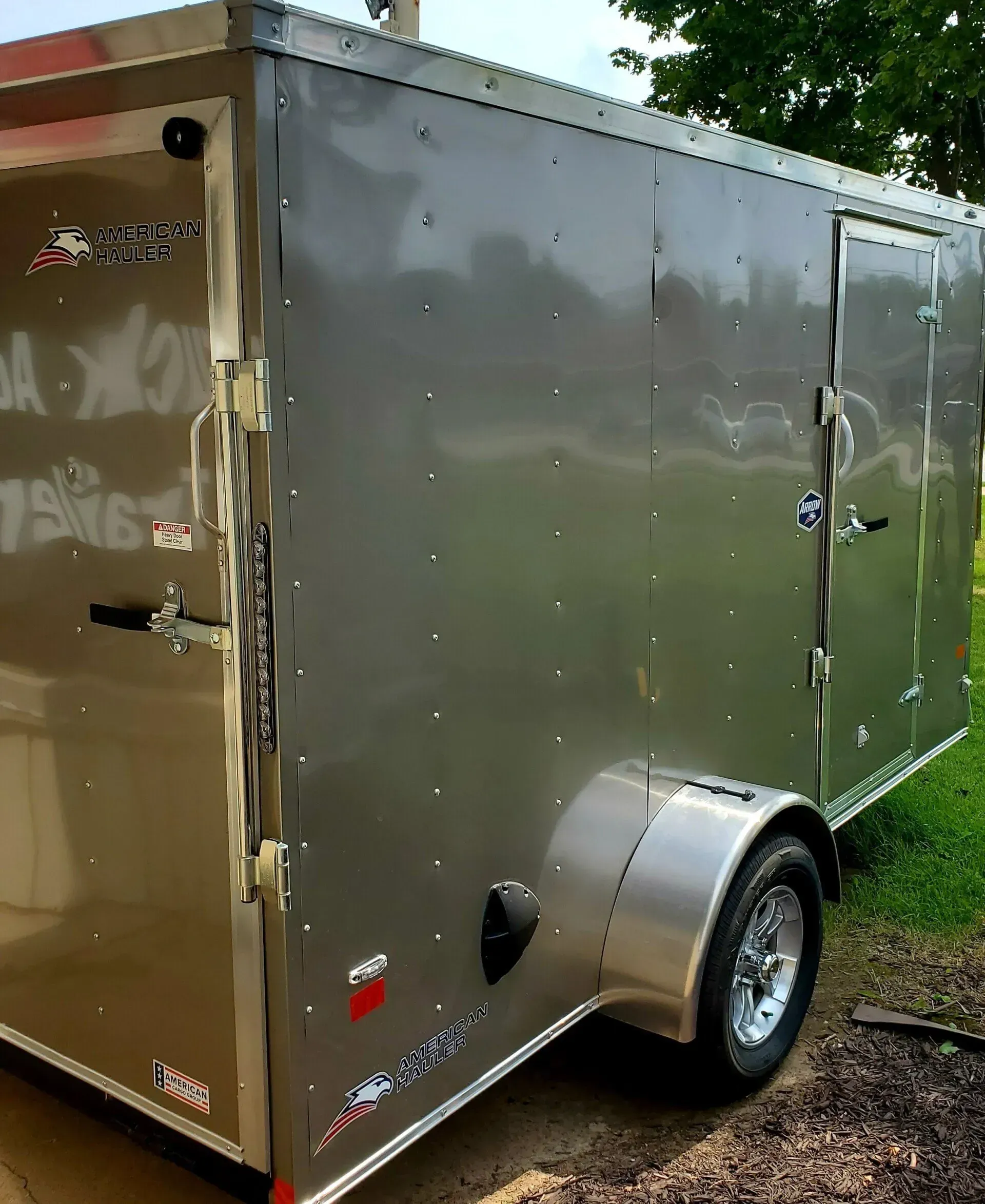 Silver enclosed cargo trailer parked outdoors on grass, with a side door and single wheel visible.