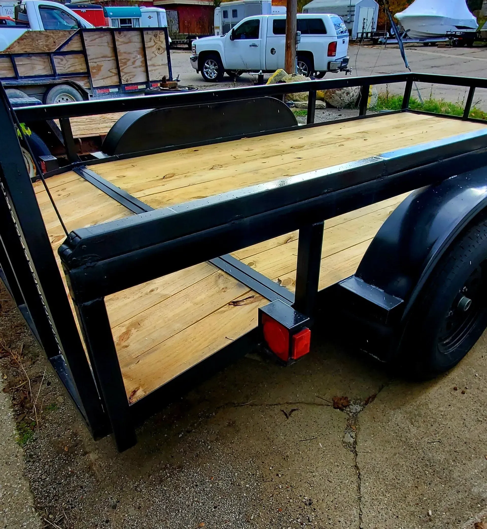 Black utility trailer with plywood floor and single wheel on wet gravel at a campsite.