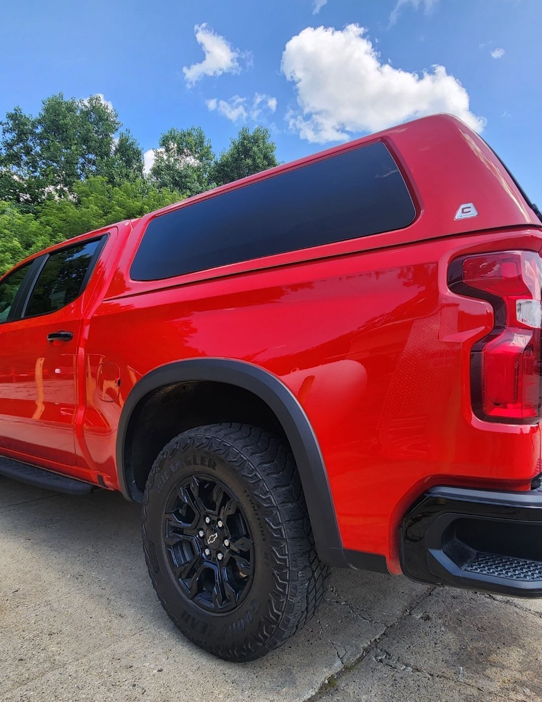 Red pickup truck with black canopy and off-road tires parked on a driveway under a blue sky.