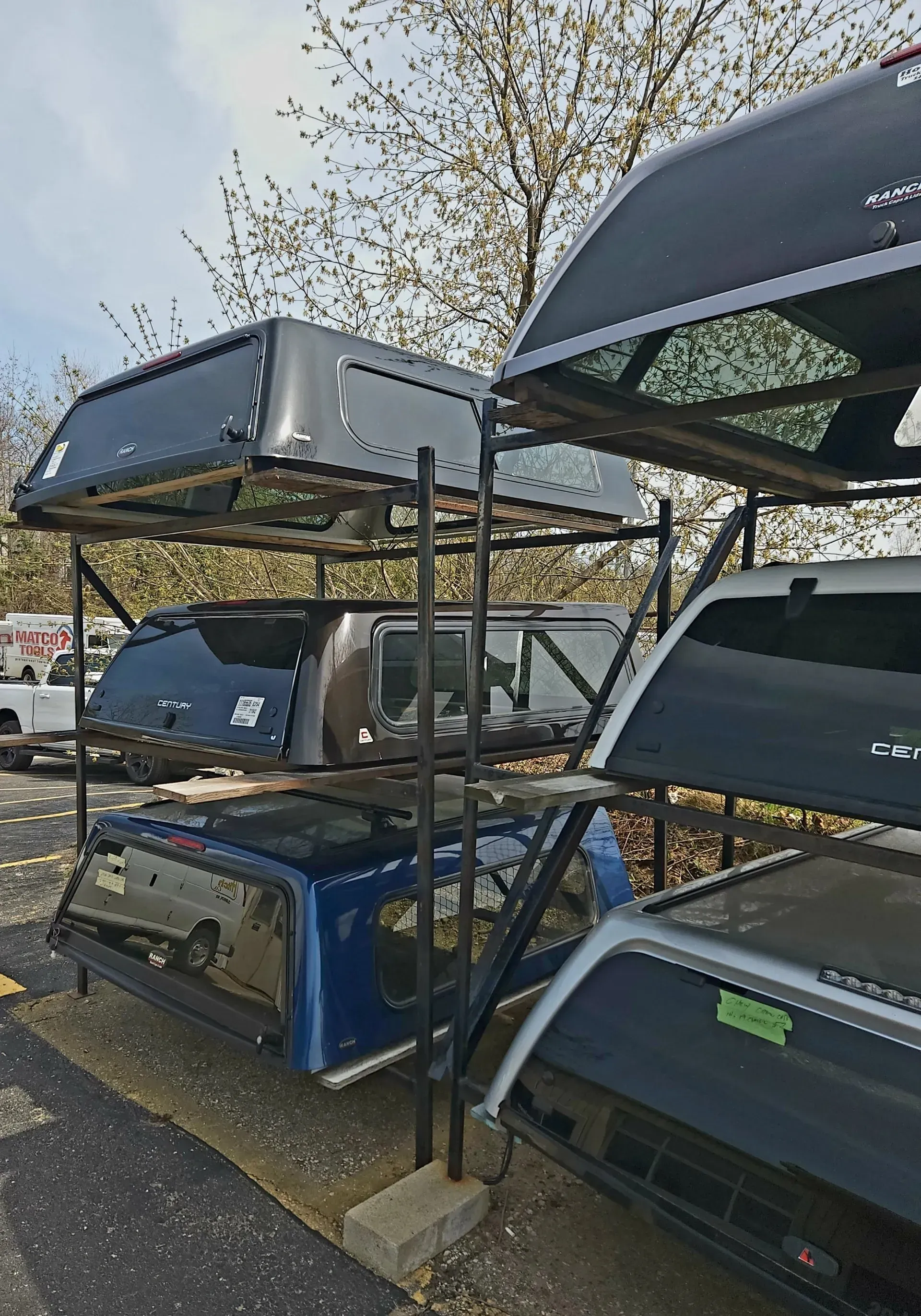 Stacked truck cap shells on outdoor metal racks, with black, blue, and gray covers in a parking lot.