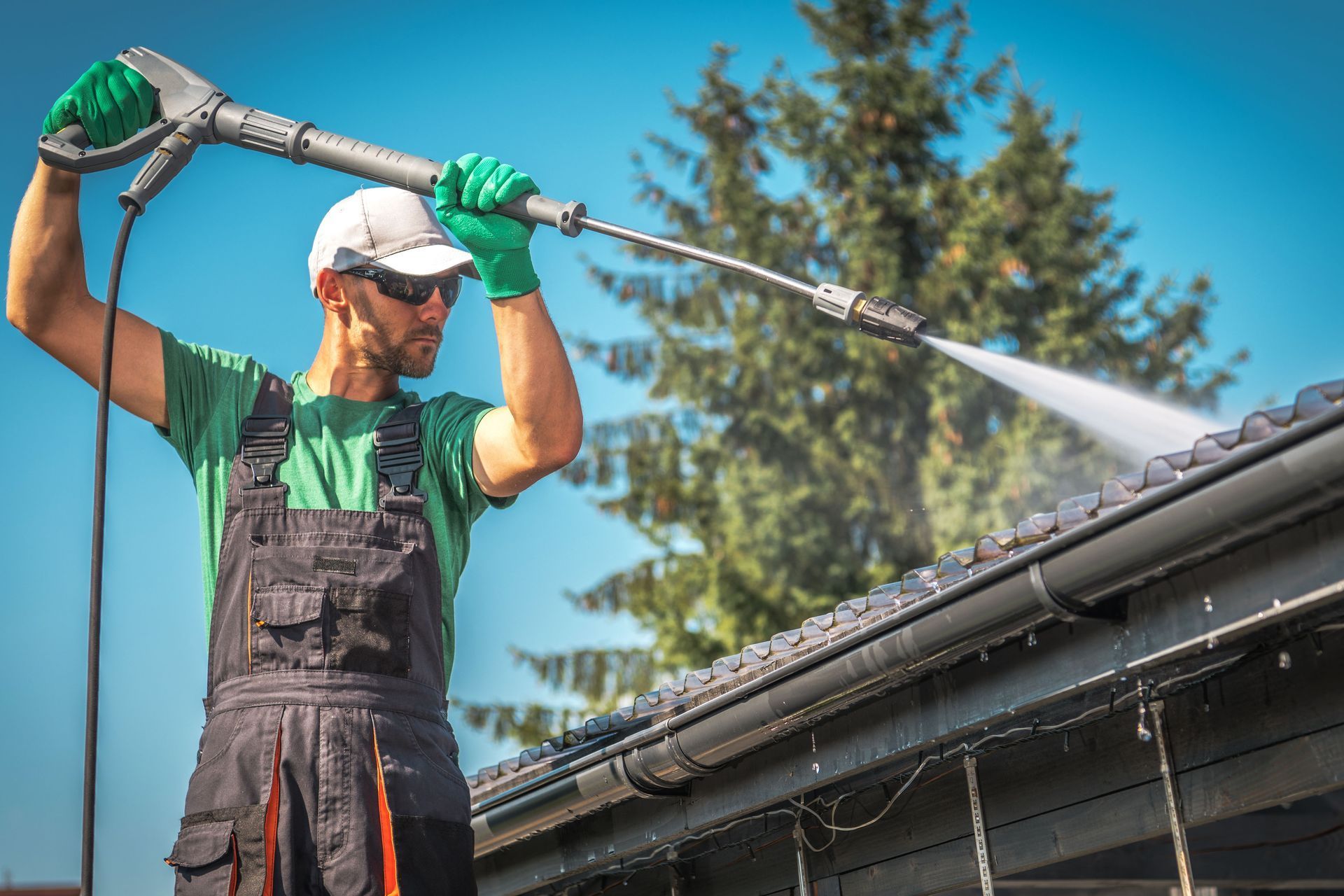 A person in green gloves, a t-shirt, and work overalls uses a pressure washer to clean the edge of a roof under a blue sky.